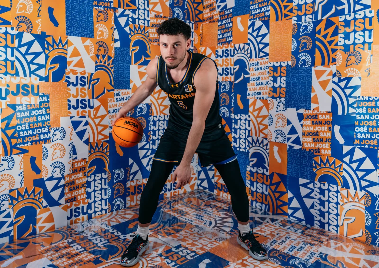 Luke Howard holding a basketball in a black San José State jersey against a blue-and-gold backdrop